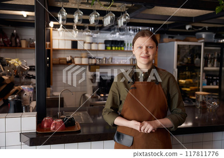 Portrait of smiling woman barista wearing apron standing in coffee shop Portrait of smiling woman barista wearing apron standing in coffee shop 117811776