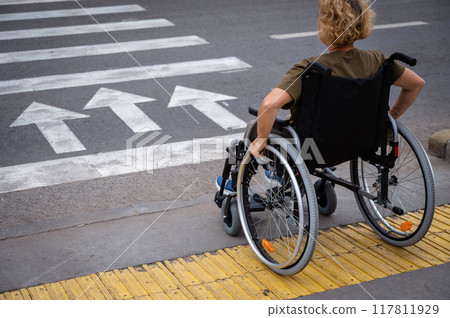 Rear view of an elderly woman on a wheelchair going to a pedestrian crossing. Rear view of an elderly woman on a wheelchair going to a pedestrian crossing. 117811929