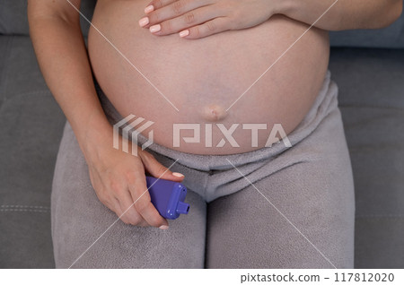 A pregnant woman smokes a vape while sitting on the couch. Close-up of the abdomen. 117812020