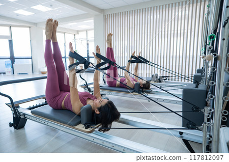 Two young asian women doing pilates exercises on a reformer.  117812097