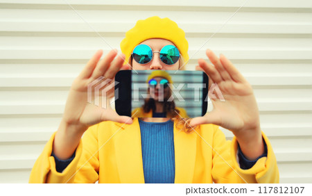 Close up modern stylish happy young woman taking selfie with phone in yellow autumn french beret hat Close up modern stylish happy young woman taking selfie with phone in yellow autumn french beret hat 117812107