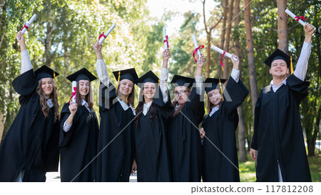 Row of young students in graduation gowns outdoors showing off their diplomas.  117812208