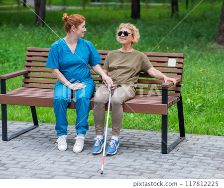 A nurse and an elderly blind woman are sitting on a bench in the park. A nurse and an elderly blind woman are sitting on a bench in the park. 117812225