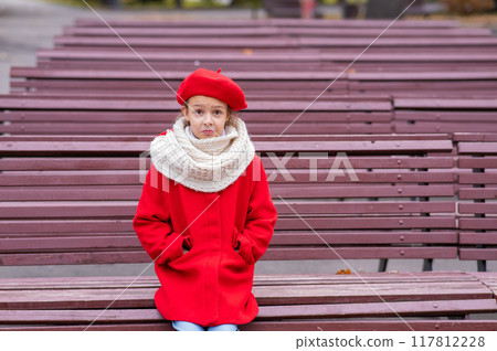 A sad Caucasian girl in a red coat and a beret sits alone on a bench.  117812228