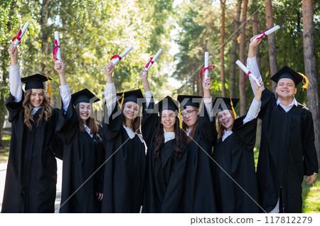 Row of young students in graduation gowns outdoors showing off their diplomas.  117812279