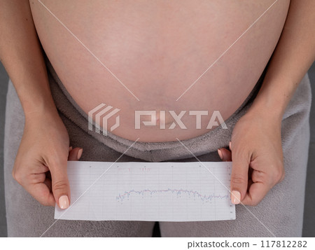 A pregnant woman looks at the results of cardiotocography while sitting on the couch. Close up of the belly. 117812282
