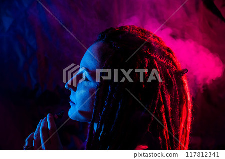 Profile of caucasian girl with dreadlocks smokes a vape in red blue light. Profile of caucasian girl with dreadlocks smokes a vape in red blue light. 117812341