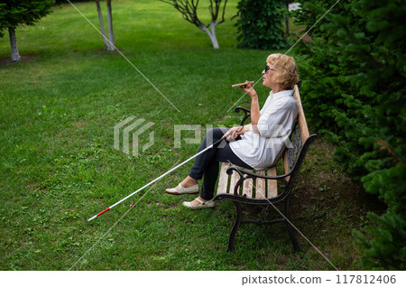 Elderly blind woman talking on a smartphone while sitting on a bench in the park. 117812406