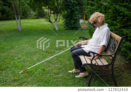 An elderly blind woman sits on a bench in the park. 117812410