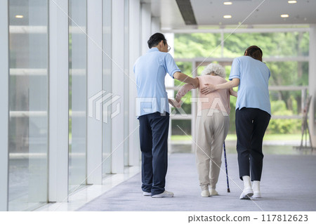 Care staff supporting a senior woman walking with a cane, back view 117812623