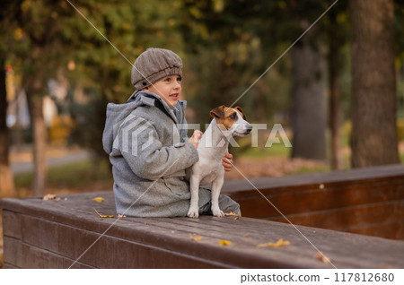 Caucasian boy sits on a bench with a dog Jack Russell Terrier for a walk in the autumn park. Caucasian boy sits on a bench with a dog Jack Russell Terrier for a walk in the autumn park. 117812680