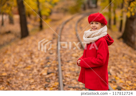 Caucasian girl in a red coat and beret walks along the railway tracks in the park in autumn. 117812724