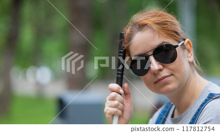 Blind red-haired woman sitting outdoors leaning on a cane.  117812754