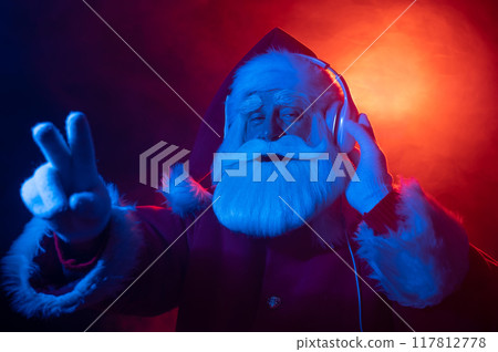 Santa Claus shows 2 fingers listening to music on headphones at a party. Blue red neon light and smoke. 117812778
