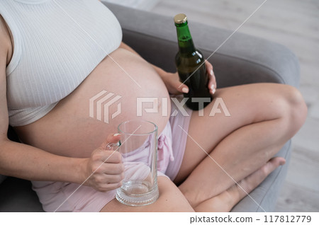 Faceless pregnant woman sitting on the sofa and holding a bottle of beer and a glass.  117812779
