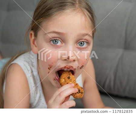 A cute little girl smeared in chocolate eats cookies while lying on the sofa. A cute little girl smeared in chocolate eats cookies while lying on the sofa. 117812835