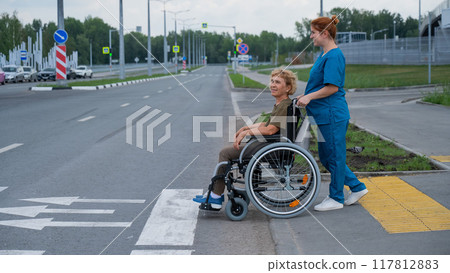 Red-haired nurse pushing an elderly woman in a wheelchair across the road. Red-haired nurse pushing an elderly woman in a wheelchair across the road. 117812883