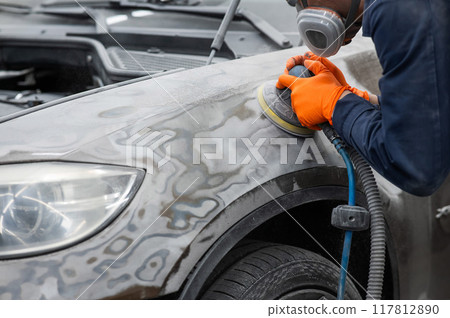 A mechanic sands the putty on a car body with a machine. Repair after an accident. A mechanic sands the putty on a car body with a machine. Repair after an accident. 117812890