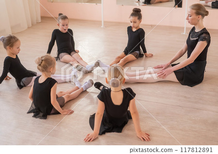 Caucasian woman and five little girls sit in a circle and do stretching at a ballet school.  117812891