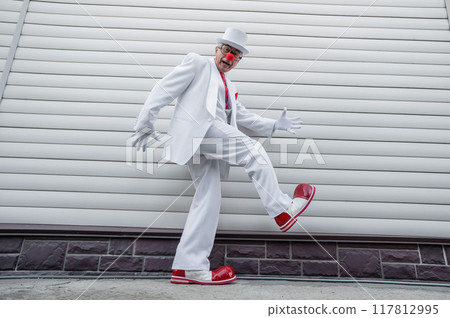 An elderly man in a white suit, huge boots and a clown nose walks funny.  117812995