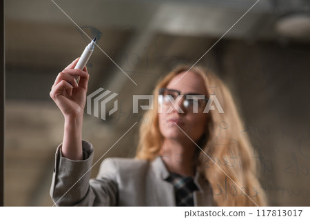 Caucasian woman with glasses writes text in English on a glass wall. Caucasian woman with glasses writes text in English on a glass wall. 117813017