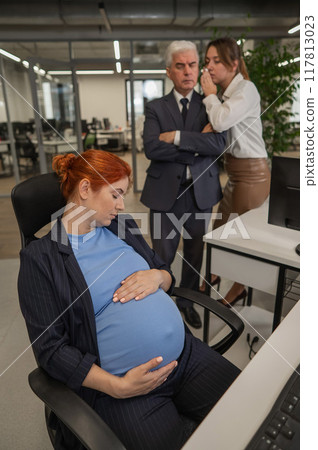 Elderly Caucasian man and woman gossiping behind their sleeping pregnant colleague in the office. Vertical photo.  117813023