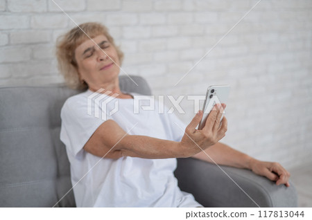 An elderly Caucasian woman suffers from farsightedness and tries to read a message on a smartphone with her arm outstretched.  117813044