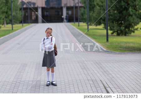 A sad caucasian girl in uniform and with a backpack holds her arms crossed on her chest stands outdoors after school. A sad caucasian girl in uniform and with a backpack holds her arms crossed on her chest stands outdoors after school. 117813073