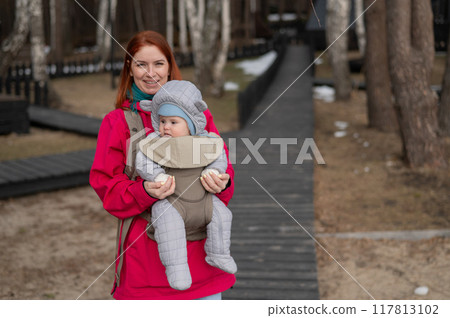 A Caucasian woman walks with her son in an ergo backpack in the countryside. A Caucasian woman walks with her son in an ergo backpack in the countryside. 117813102