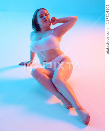 Portrait of young beautiful woman sitting confidently in textured swimwear and posing, showing self-love against blue background background in neon light Portrait of young beautiful woman sitting confidently in textured swimwear and posing, showing self-love against blue background background in neon light 117814101