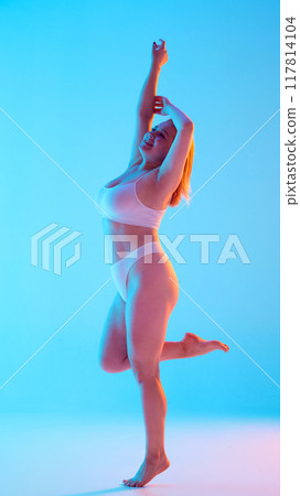 Full-length image of young attractive woman with plus-size body posing in white underwear against blue background background in neon light 117814104