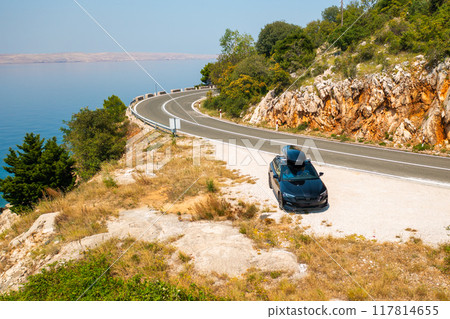 Black car with a roof box, parked at the edge of a narrow, winding road on a rugged cliff.  117814655