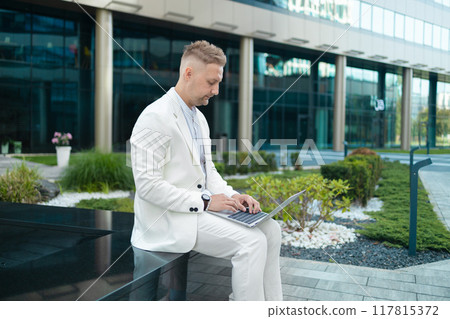 Focused young businessman corporate worker investor sitting near business centre waiting, working remote on laptop Focused young businessman corporate worker investor sitting near business centre waiting, working remote on laptop 117815372