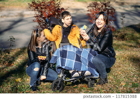 Mother and sibling engaging with child in wheelchair during outdoor bubble play, family support, special needs care, nurturing joy, disability inclusion. Mother and sibling engaging with child in wheelchair during outdoor bubble play, family support, special needs care, nurturing joy, disability inclusion. 117815388