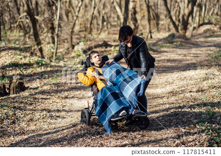 Mother adjusting blanket for child in wheelchair during outdoor walk in forest, caregiving, special needs support, family outings, disability inclusion. 117815411