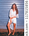 Summery boho look: young woman in white lace in front of a blue wooden wall 117815856