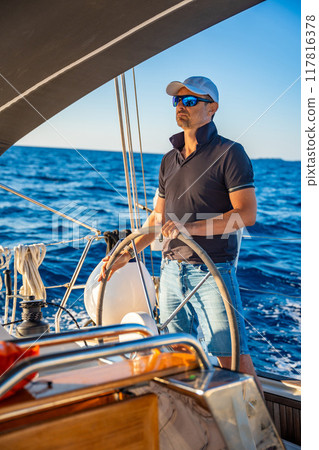Young man captain stands at the helm and controls a sailboat during a journey by sea Young man captain stands at the helm and controls a sailboat during a journey by sea 117816378
