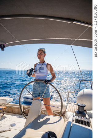 Young female captain on the yacht looking through binoculars during sailing boat control. Travel and active life. Yachting 117816380