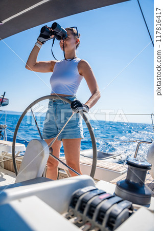 Young female captain on the yacht looking through binoculars during sailing boat control. Travel and active life. Yachting 117816417