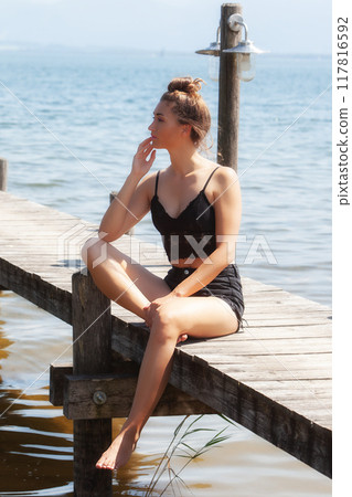 Thoughtful pause by the lake: woman in a black lace outfit on a jetty Thoughtful pause by the lake: woman in a black lace outfit on a jetty 117816592