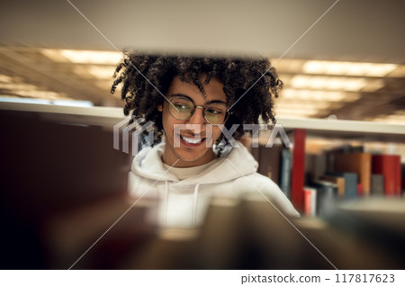 Cheerful smiling male student with curly hair standing with near bookshelf Cheerful smiling male student with curly hair standing with near bookshelf 117817623