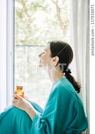 Woman in blue sitting by the window holding candle, practicing autumn fall self-care, mindfulness, and relaxation. 117818071