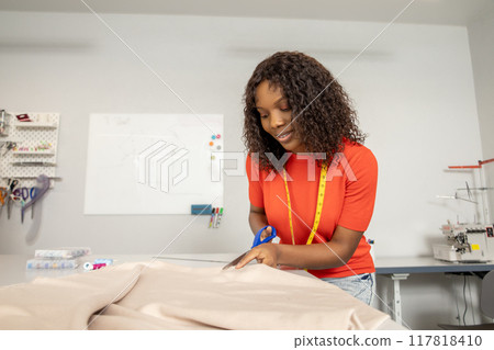 African American woman dressmaker cutting dress fabric on sketch line with scissors African American woman dressmaker cutting dress fabric on sketch line with scissors 117818410