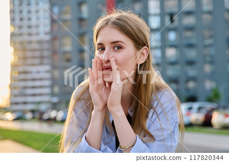 Young cheerful woman whispering secret, holding hands near mouth, outdoor 117820344