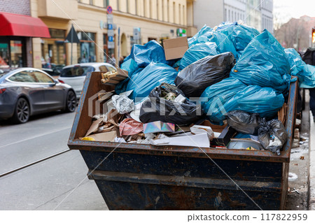 Big metal overloaded dumpster skip container full of plastic trash litter bags near residential building at Berlin city Mitte downtown or suburban area. Non-recyclable sorting garbage disposal 117822959
