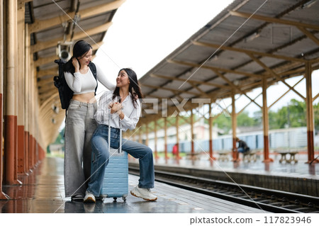 Two women are sitting on a train platform with their luggage Two women are sitting on a train platform with their luggage 117823946