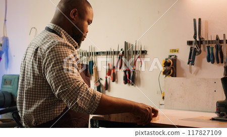BIPOC man inspecting wood in assembling shop, putting protection equipment on and picking tool from rack. African american cabinetmaker picking lumber block, preparing to do piling on it, camera A 117827109