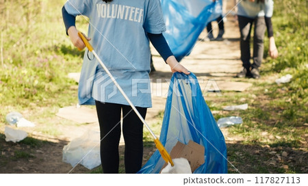 Young child using tongs tool to grab and pick up trash from the woods, collecting rubbish from a natural habitat. Little girl growing up with environmental care and recycling plastic waste. Camera A. 117827113