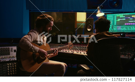 Performer songwriter singing new tracks on his electro acoustic guitar and recording new music in post production. Musician working in collaboration with audio engineer in studio. Camera A. 117827114