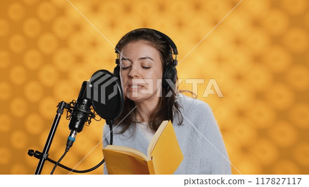 Voice actor reading book, recording for audiobook production using microphone and headset, studio background. Person using mic and headphones to produce digital recording of novel, camera A Voice actor reading book, recording for audiobook production using microphone and headset, studio background. Person using mic and headphones to produce digital recording of novel, camera A 117827117